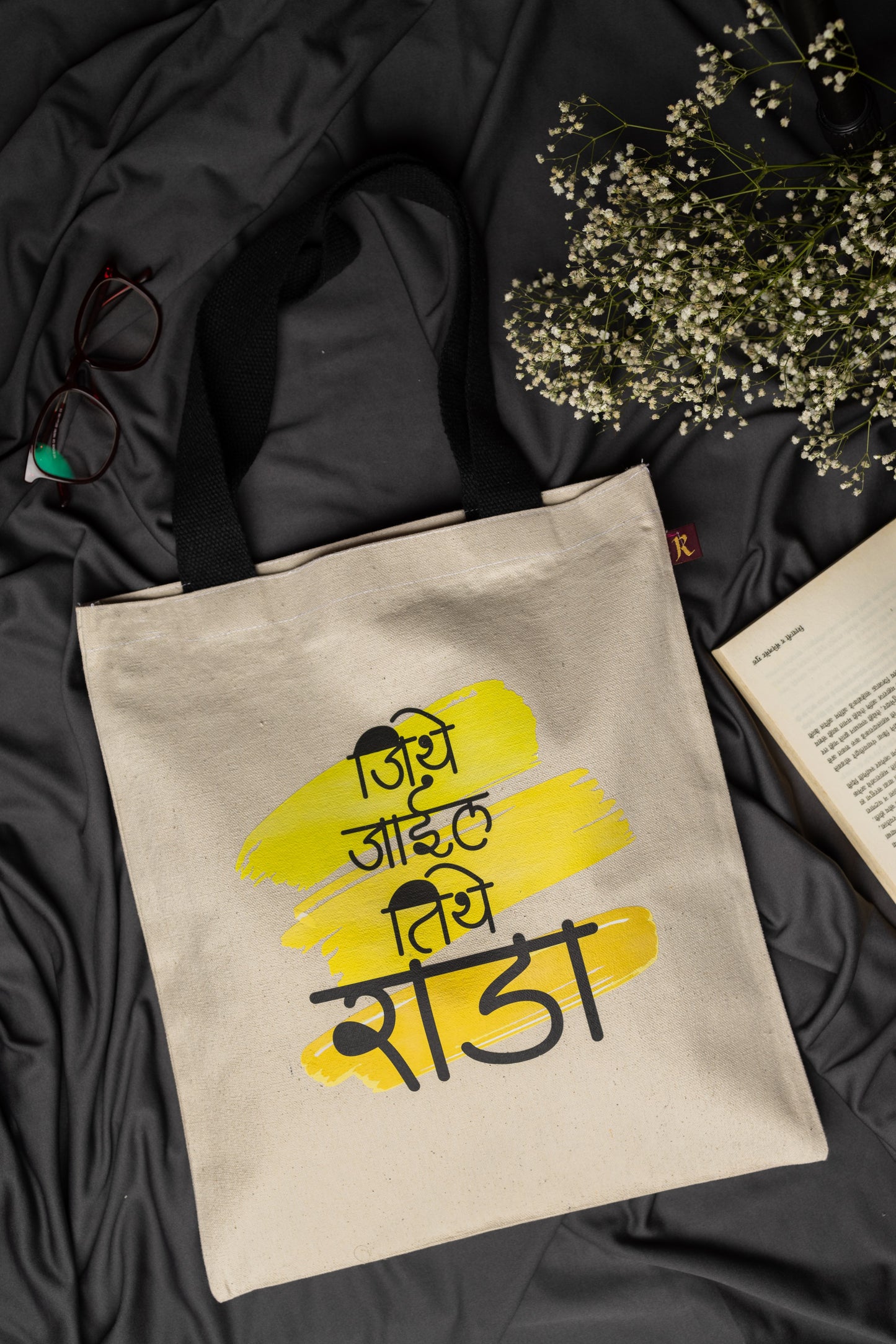 Beige tote bag with yellow and black text on a dark fabric background with flowers and glasses.