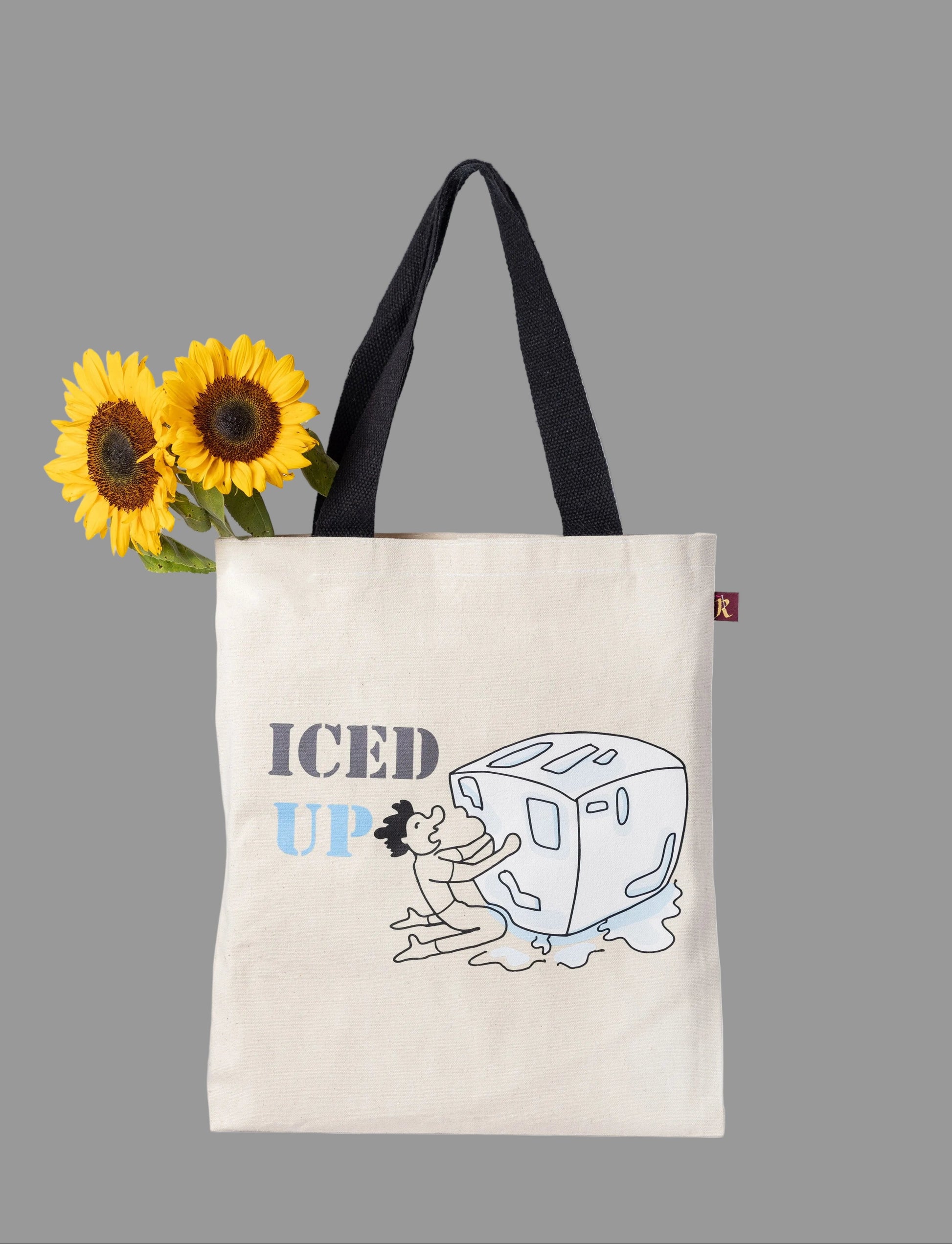 A off white cotton tote bag with 'ICED UP' printed on it, featuring a whimsical illustration of a character hugging a giant ice cube. The bag has black handles.