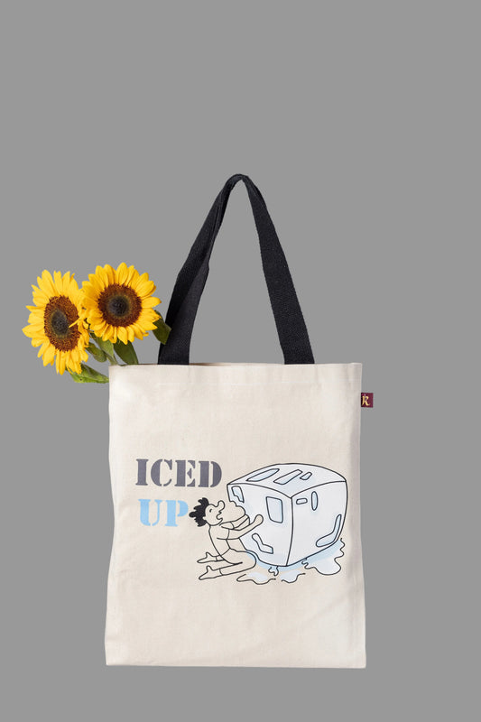 A off white cotton tote bag with 'ICED UP' printed on it, featuring a whimsical illustration of a character hugging a giant ice cube. The bag has black handles.