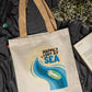 Beige tote bag with 'Happily Lost at Sea' design on a dark fabric background