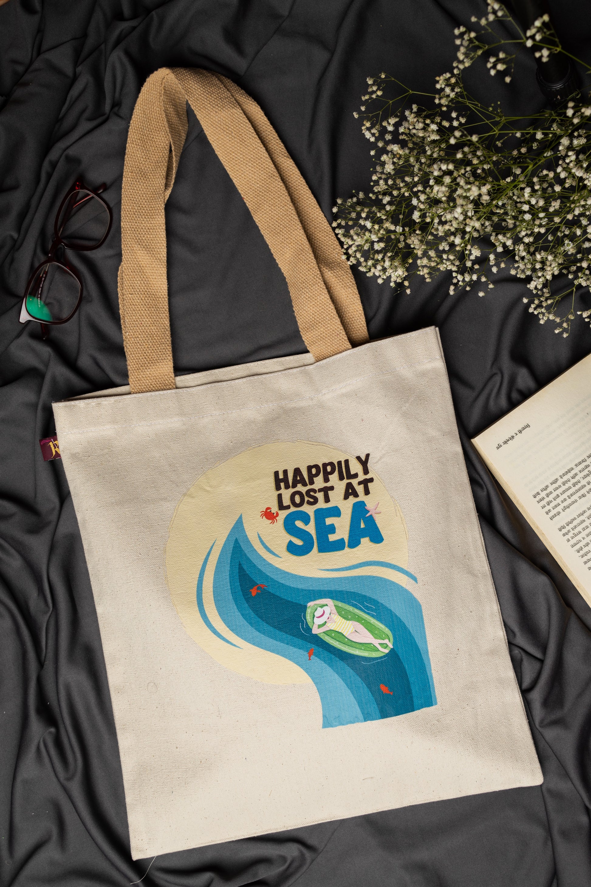 Beige tote bag with 'Happily Lost at Sea' design on a dark fabric background