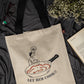 Beige tote bag with a graphic design and text 'LET HIM COOK!' on a dark fabric background.