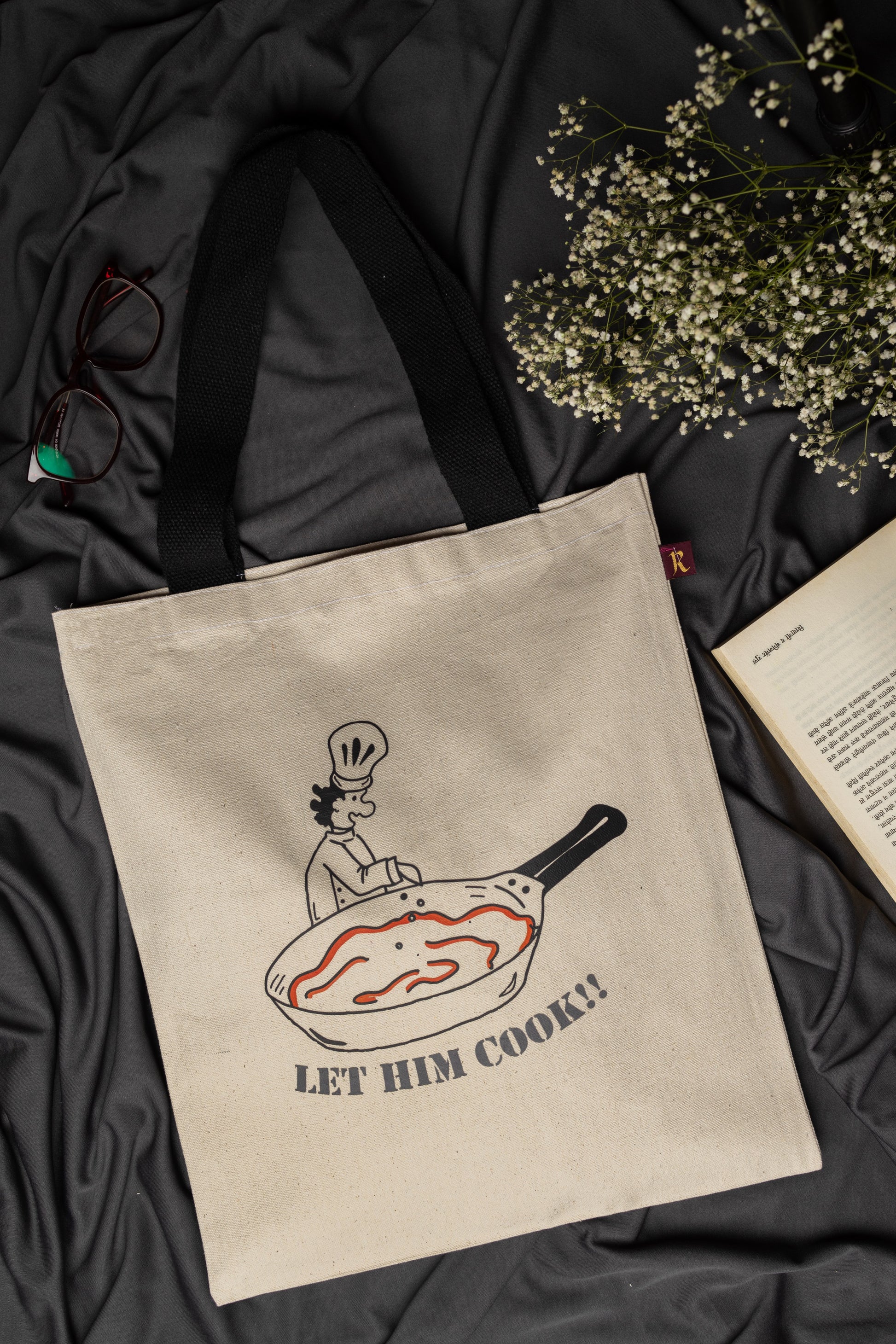 Beige tote bag with a graphic design and text 'LET HIM COOK!' on a dark fabric background.
