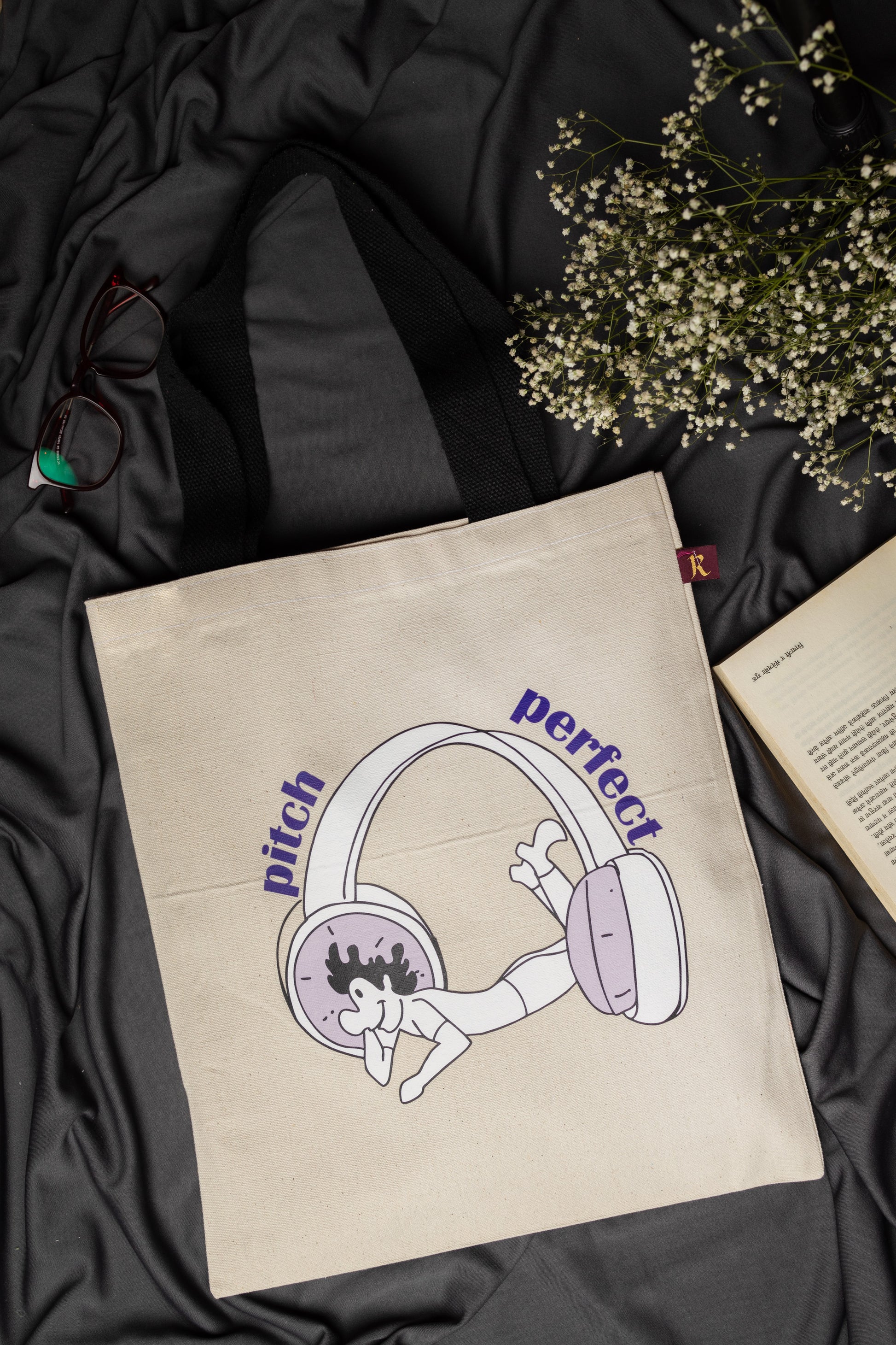 Beige tote bag with headphone illustration and text on a dark fabric background
