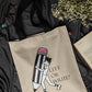 Beige tote bag with a design of a person holding a pencil and text 'LEFT OR WRITE?' on a dark fabric background.