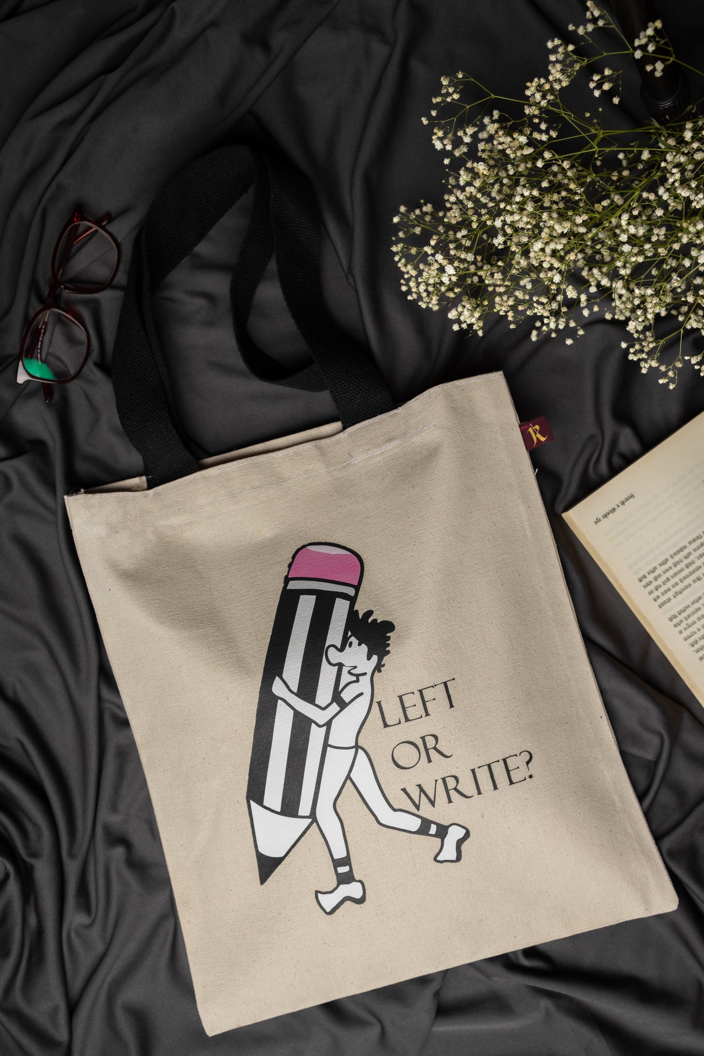 Beige tote bag with a design of a person holding a pencil and text 'LEFT OR WRITE?' on a dark fabric background.
