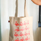 A beige cotton tote bag with a printed pattern of pink flamingos on it, held up against a light background.