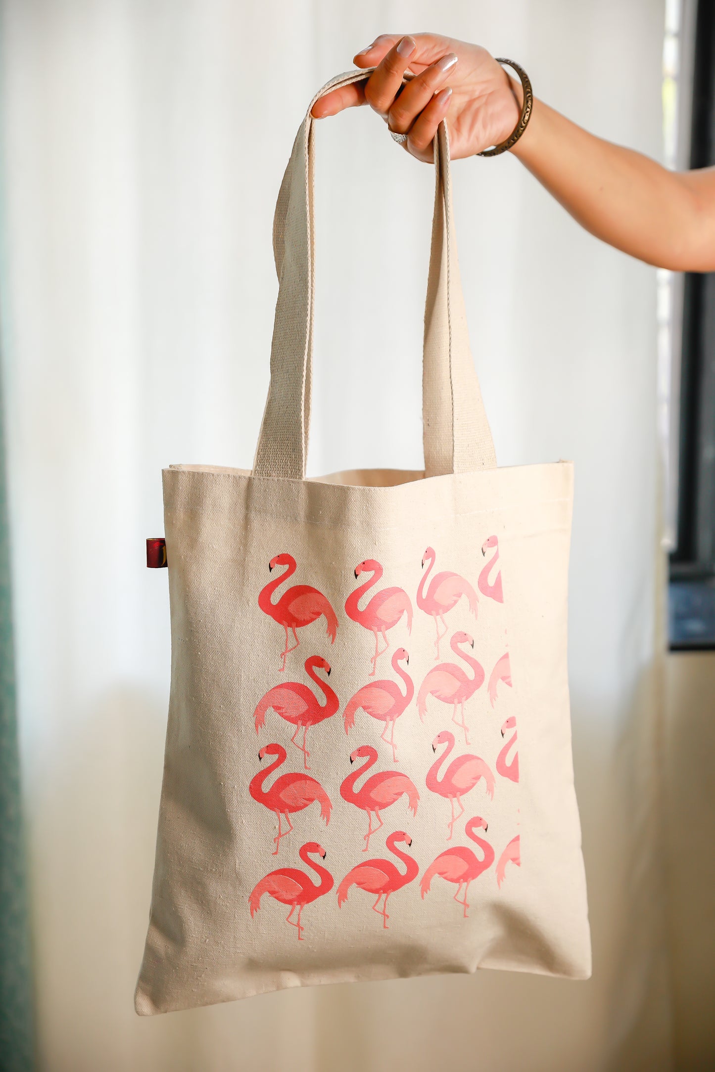 A beige cotton tote bag with a printed pattern of pink flamingos on it, held up against a light background.