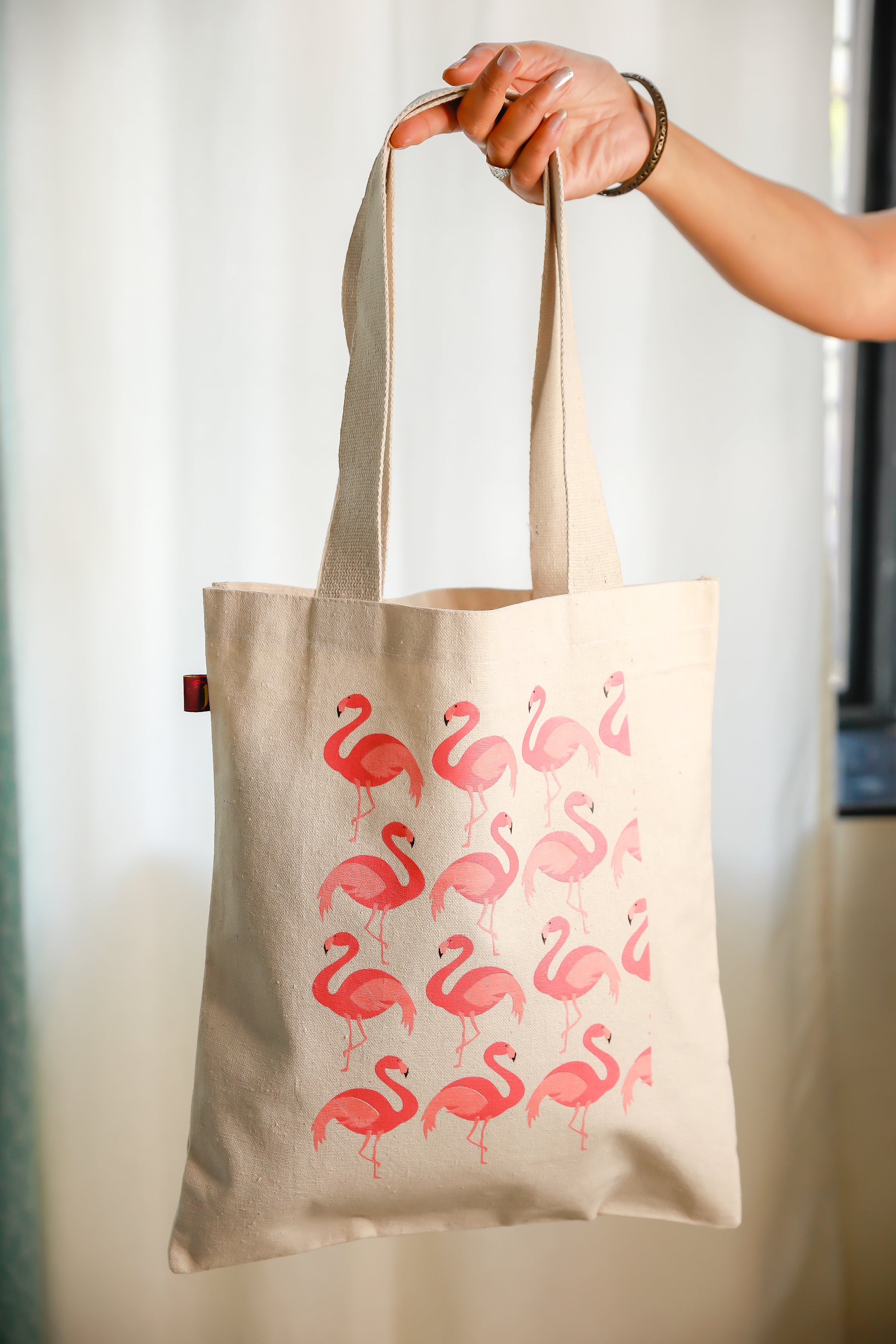 A beige cotton tote bag with a printed pattern of pink flamingos on it, held up against a light background.