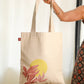 A medium-sized flat tote bag in beige, featuring a printed design with a sun and three leaves. The bag is being held by a person.