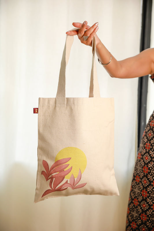 A medium-sized flat tote bag in beige, featuring a printed design with a sun and three leaves. The bag is being held by a person.