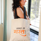 A person holding a beige flat tote bag with black straps and text printed in Marathi