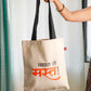 A person holding a beige flat tote bag with black straps and text printed in Marathi