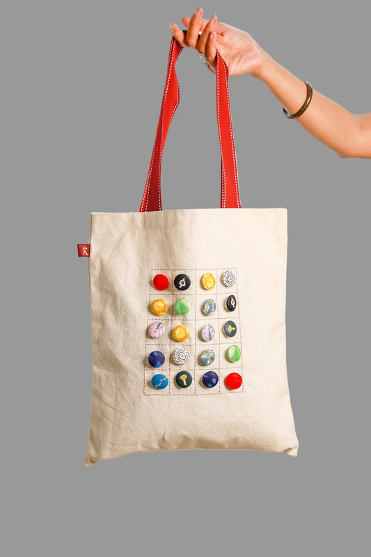 A medium-sized beige flat tote bag with a pattern of fabric buttons on the front, held up against a blurred background.
